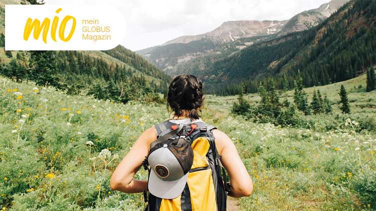 Person mit Rucksack in bergiger Landschaft mit Wiesen und Bäumen, oben links das mio mein GLOBUS Magazin-Logo in Gelb und Weiß.