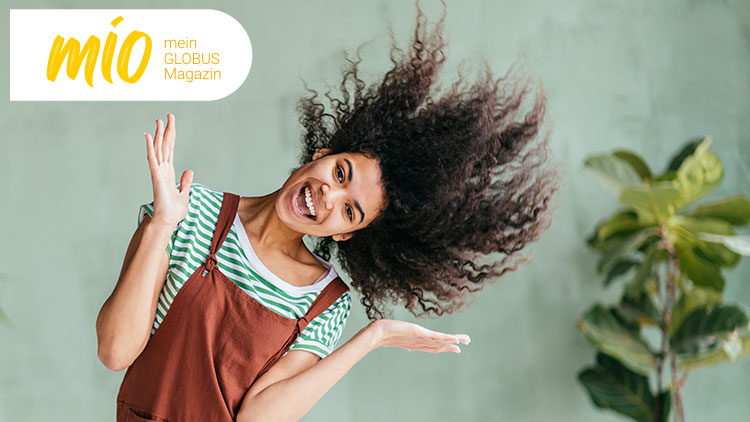 Person in braunem Latzkleid und grün-weiß gestreiftem Shirt schwingt Haare, links oben steht „mio mein GLOBUS Magazin“.