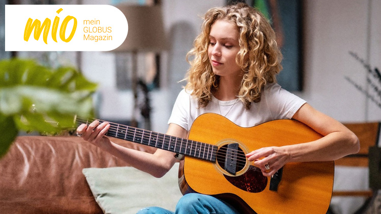 Person in weißem T-Shirt spielt Akustikgitarre, im Hintergrund Wohnzimmeratmosphäre, mio mein GLOBUS Magazin-Logo oben links.