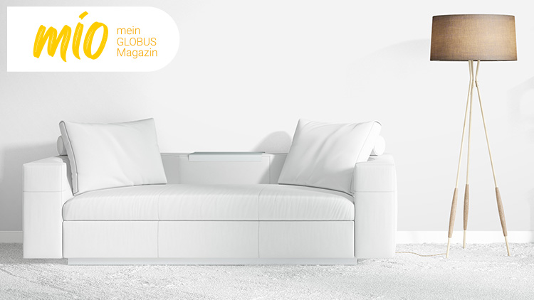 Ein weißes Sofa mit zwei Kissen, eine Stehlampe und ein Regal mit Pflanzen, rechts oben steht „mio, mein GLOBUS Magazin“.