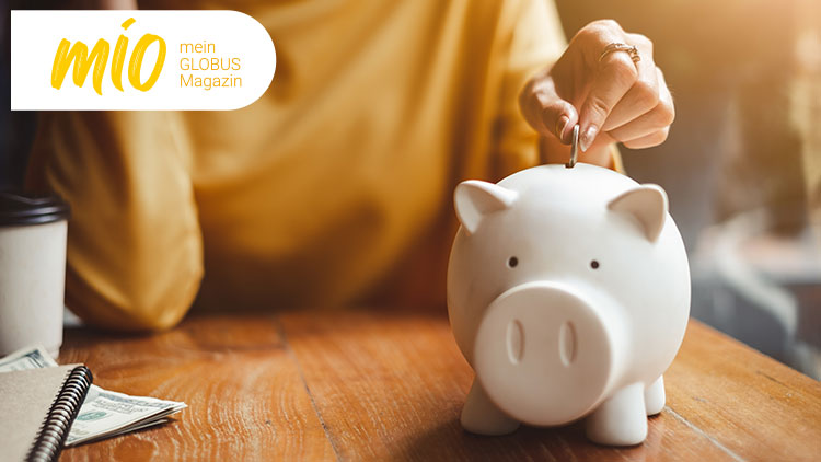 Eine Hand steckt eine Münze in ein weißes Sparschwein, daneben liegt Geld und ein Getränkebecher, oben steht „mio mein GLOBUS Magazin“.
