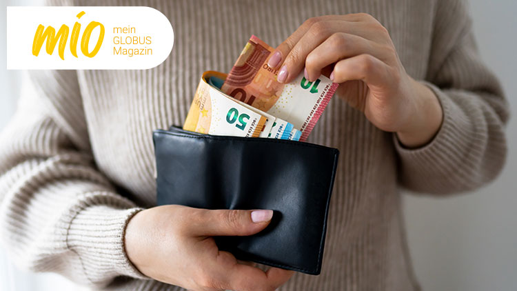 Eine Hand steckt Geldscheine in eine schwarze Geldbörse, oben links steht „mio mein GLOBUS Magazin“ auf weißem Hintergrund.