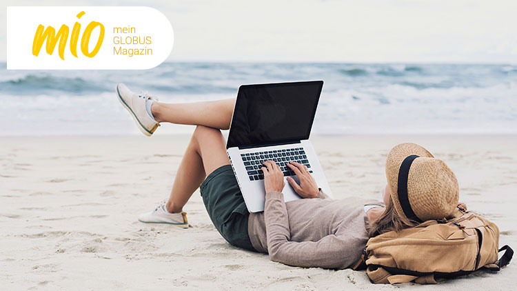 Person liegt entspannt am Strand, arbeitet auf einem Laptop, daneben liegt ein Rucksack und ein Hut, oben links Text „mio mein GLOBUS Magazin“.