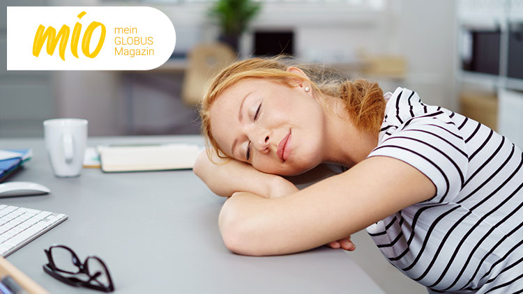 Eine Person mit gestreiftem Oberteil liegt mit dem Kopf auf einem Tisch, davor Tastatur, Maus und Ordner, links Text „mio mein GLOBUS Magazin“.