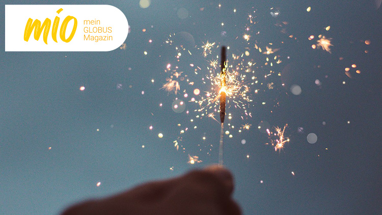 Ein brennendes Wunderkerzenlicht vor unscharfem Hintergrund, oben links steht mio mein GLOBUS Magazin in weiß-gelbem Logo.