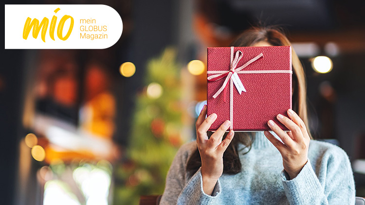 Person hält ein rotes Geschenk mit weißer Schleife vor ihrem Gesicht, links oben steht „mio mein GLOBUS Magazin“ auf weißem Hintergrund.