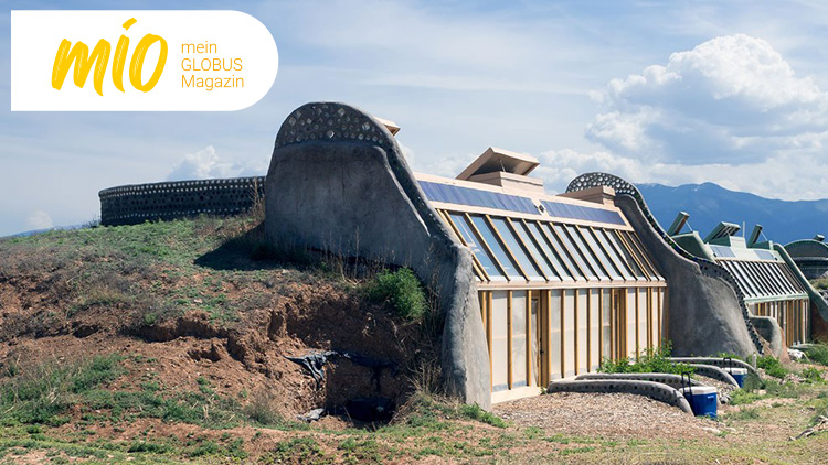 Ein Gebäude mit gewölbten Wänden, begrüntem Dach und großen Fenstern steht in einer Landschaft mit Bergen im Hintergrund, mio mein GLOBUS Magazin.