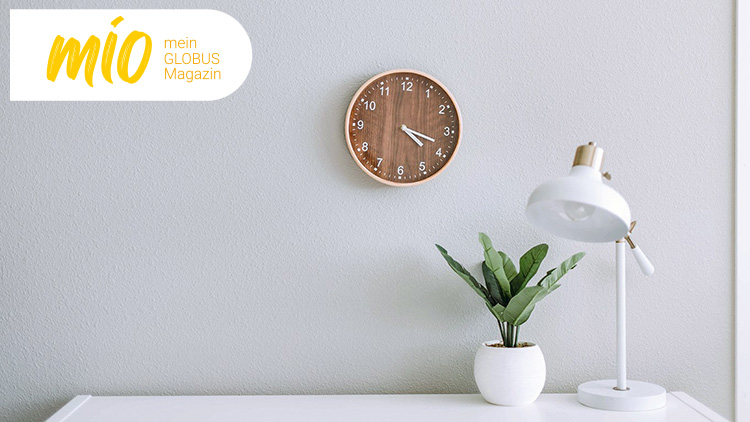 An weißer Wand eine runde Holzuhr, darunter eine weiße Lampe mit goldenen Details und eine grüne Pflanze, oben Schriftzug „mio mein GLOBUS Magazin“.