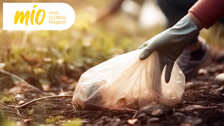 Eine behandschuhte Hand hebt eine Plastiktüte vom Boden auf, umgeben von unscharfer Vegetation, oben steht „mio mein GLOBUS Magazin“.