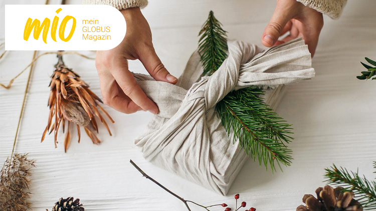 Hände wickeln ein Geschenk in beigen Stoff mit Tannenzweigen, links oben steht „mio mein GLOBUS Magazin“ auf weißem Hintergrund.
