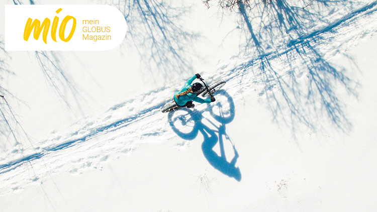 Eine Person fährt mit einem Fahrrad über Schnee, wirft einen langen Schatten, oben links steht „mio mein GLOBUS Magazin“.