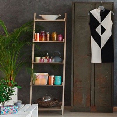 Ein Holztisch mit Pflanzen steht vor einem Regal mit Gläsern und Keramik, daneben ein Schrank mit einem schwarz-weißen Kleid.