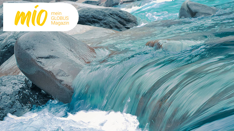 Ein klarer Fluss fließt mit türkisfarbenem Wasser über glatte graue Felsen, oben links steht „mio mein GLOBUS Magazin“.