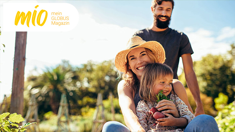 Person mit Strohhut hält Gemüse, im Hintergrund verschwommene grüne Gartenlandschaft und das mio mein GLOBUS Magazin-Logo.