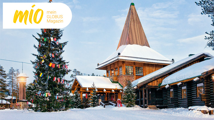 Verschneite Holzhäuser mit Tannenbäumen, beleuchtetem Weihnachtsbaum und dem mio mein GLOBUS Magazin-Logo links oben.