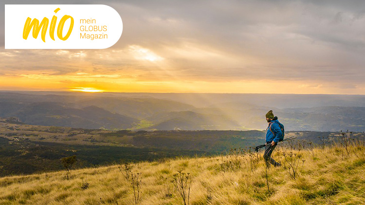 Person in Outdoor-Kleidung läuft mit Rucksack und Stöcken über eine hügelige Wiese bei Sonnenuntergang, Text: mio mein GLOBUS Magazin.