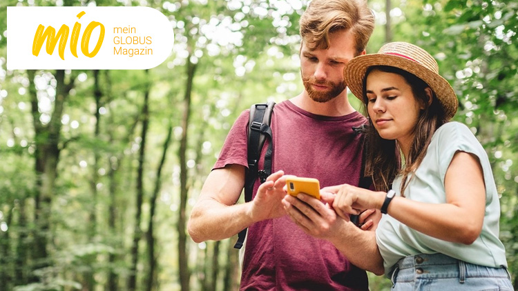 Zwei Personen in Freizeitkleidung stehen im Wald, eine Person hält ein gelbes Smartphone, mio mein GLOBUS Magazin-Logo oben links.