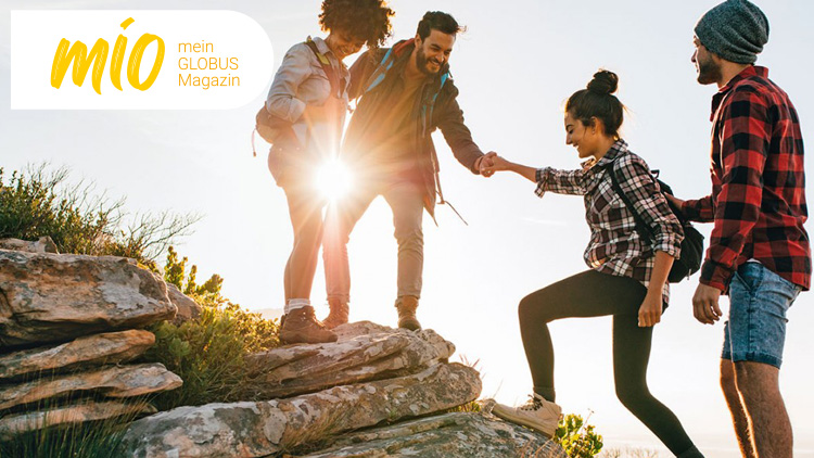 Vier Personen wandern auf einem Felsen, eine Person hilft einer anderen nach oben, links steht das mio mein GLOBUS Magazin-Logo.