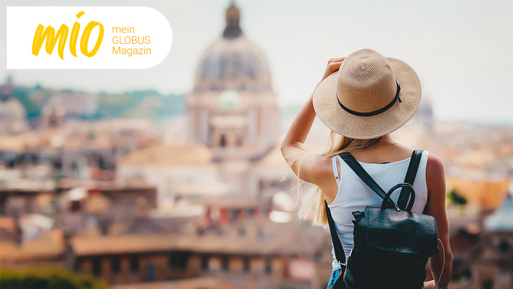 Person mit Strohhut und schwarzem Rucksack blickt auf eine Stadt mit historischen Gebäuden, oben links steht mio mein GLOBUS Magazin.
