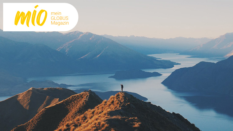 Berglandschaft mit Person auf einem Grat, im Hintergrund ein großer See und umliegende Berge, oben links mio mein GLOBUS Magazin.