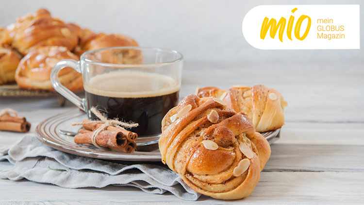 Eine Tasse schwarzer Kaffee und zwei Zimtschnecken mit Mandeln liegen auf einem Teller, daneben steht „mio mein GLOBUS Magazin“.