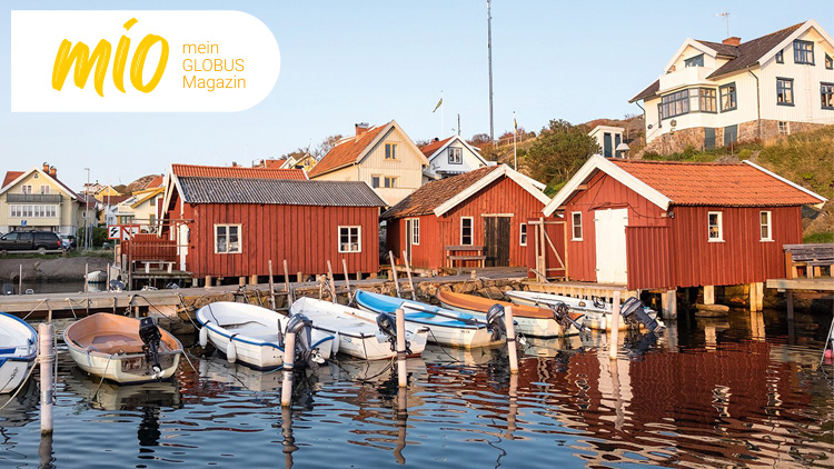 Rote Holzhäuser stehen auf Stelzen am Wasser, davor liegen kleine Boote, oben links steht „mio mein GLOBUS Magazin“.
