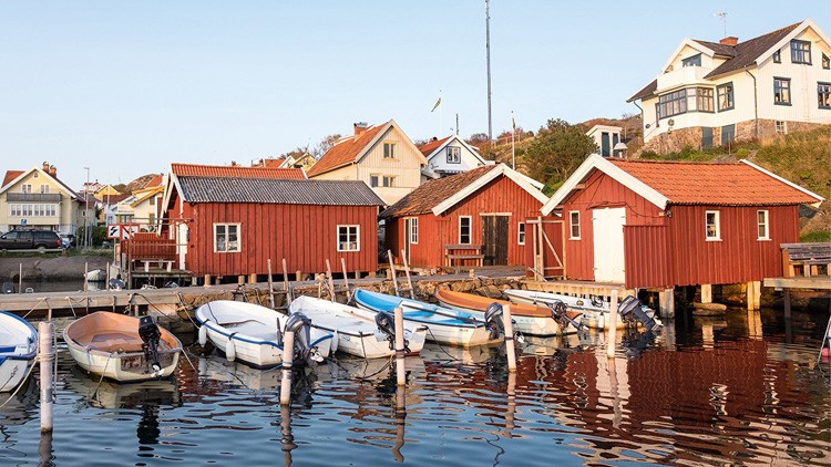 Mehrere weiße Boote liegen vor roten Holzhäusern an einem Holzsteg, dahinter stehen weiße Wohnhäuser auf einem Hügel.