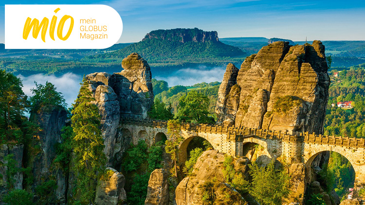 Felsformation mit Brücke in grüner Landschaft, im Hintergrund Wälder und ein Tafelberg, oben links der Schriftzug mio mein GLOBUS Magazin.