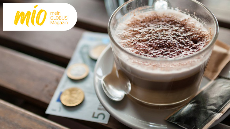 Eine durchsichtige Tasse Latte Macchiato mit Kakaopulver steht auf einem Tisch neben Münzen, einem Löffel und Servietten.