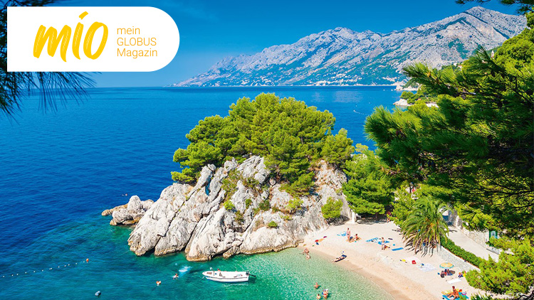 Kleine Bucht mit Sandstrand, türkisfarbenem Wasser, Booten und Badenden, umgeben von Felsen und grünen Bäumen, mio mein GLOBUS Magazin.