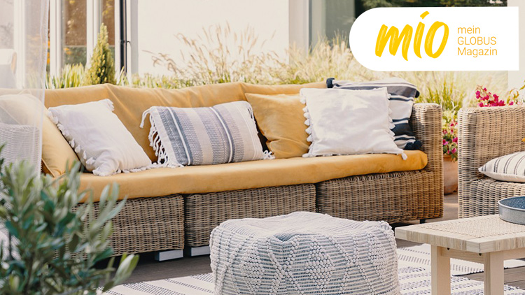 Rattan-Sitzgruppe mit gelbem Sofa und weißen Kissen, umgeben von Pflanzen, mit mio mein GLOBUS Magazin-Logo oben rechts.