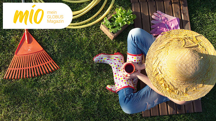 Person mit Strohhut und gepunkteten Gummistiefeln sitzt auf Rasen, hält eine rote Tasse, daneben Gartenwerkzeuge und mio mein GLOBUS Magazin.