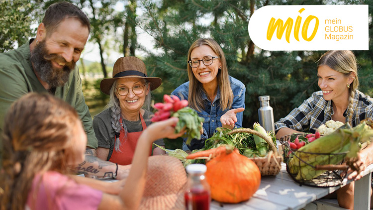 Menschen sitzen an einem Tisch im Freien mit frischem Gemüse, im Bild rechts oben steht „mio mein GLOBUS Magazin“.