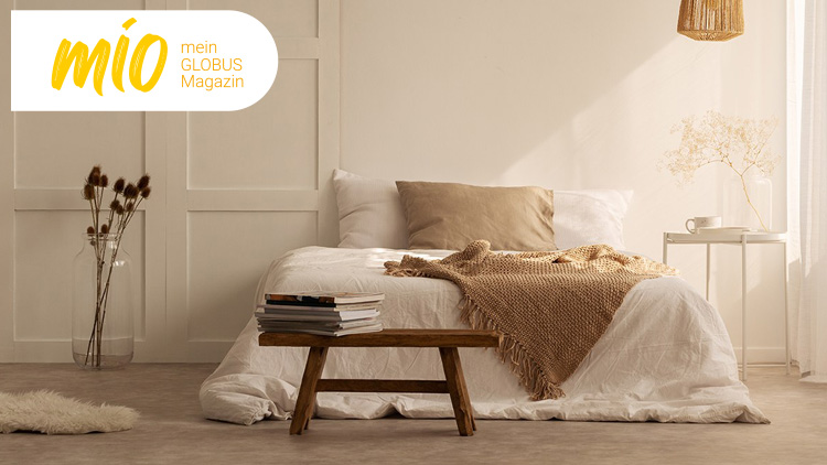Helles Schlafzimmer mit weißem Bett, beigem Kissen, brauner Decke, Holzbank, Trockenblumen und mio mein GLOBUS Magazin-Logo.