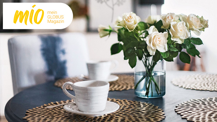 Vase mit weißen Rosen und zwei weiße Tassen auf einem gedeckten Tisch, links oben Schriftzug mio mein GLOBUS Magazin.