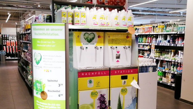 Nachfüllstation für Bio-Waschmittel mit zwei Zapfhähnen und Produkten der Marke „Planet Pure“, daneben Regale mit Reinigungsmitteln.