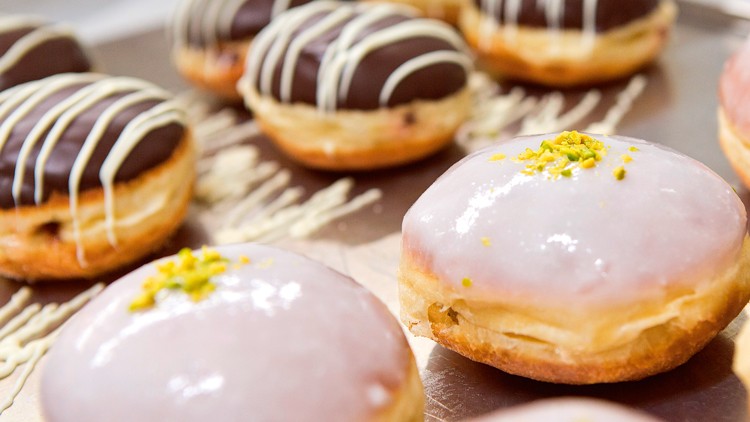 Donuts mit rosa Glasur und gehackten Pistazien sowie Donuts mit Schokoladenglasur und weißer Verzierung auf einem Tablett.