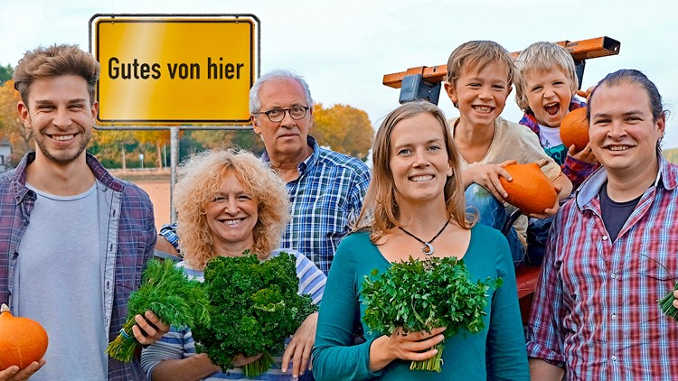 Eine Gruppe von Menschen hält Kräuter, Kürbisse und Schnittlauch vor einem gelben Schild mit der Aufschrift Gutes von hier.