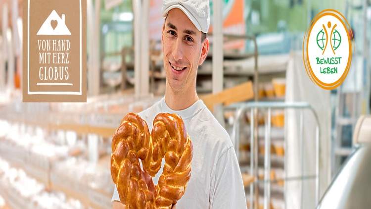 Person in weißer Kleidung hält ein herzförmiges Flechtgebäck, links ein Schild „Von Hand mit Herz GLOBUS“, rechts „Bewusst leben“.