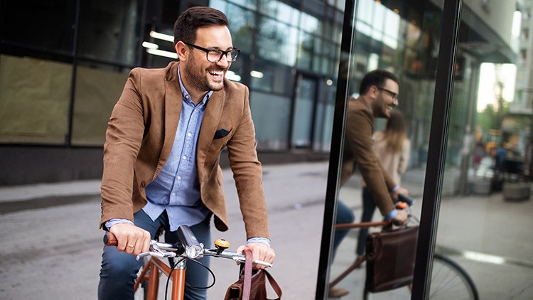 Person in brauner Jacke fährt ein Fahrrad mit Lenkerklingel, im Hintergrund sind Glasfassaden und eine Straße erkennbar.