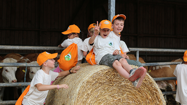Kinder in weißen T-Shirts und orangenen GLOBUS-Kappen sitzen auf einem Strohballen, im Hintergrund sind Kühe in einem Stall.
