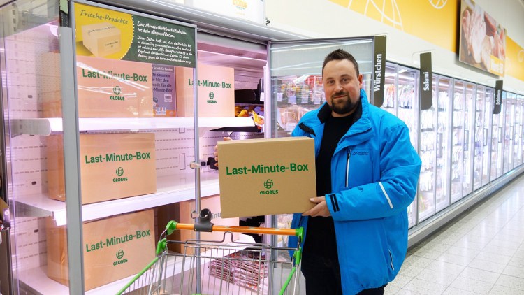 Person in blauer Jacke hält GLOBUS Last-Minute-Box vor Kühlregalen mit weiteren Last-Minute-Boxen, Einkaufswagen vorne.