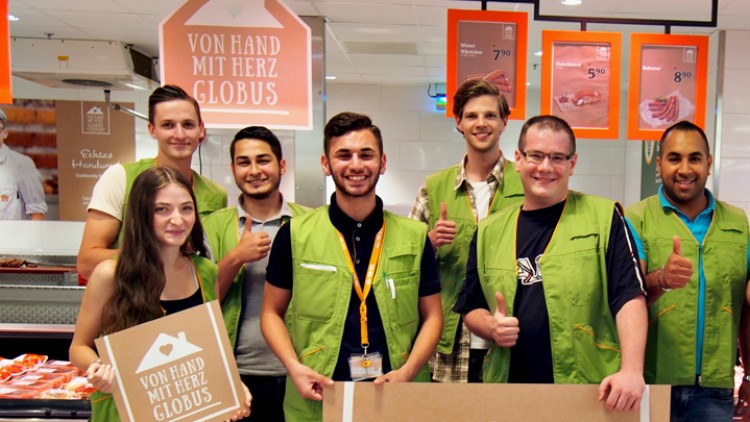 Gruppe von Personen in grünen Westen hält Schilder mit dem Text „Von Hand mit Herz GLOBUS“, im Hintergrund eine Fleischtheke.