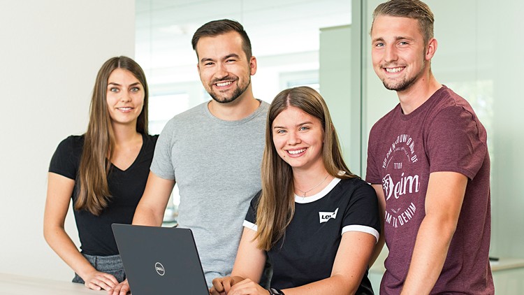 Vier Personen stehen und sitzen an einem Tisch, auf dem ein schwarzer Laptop der Marke Dell steht, hinter Glaswänden.