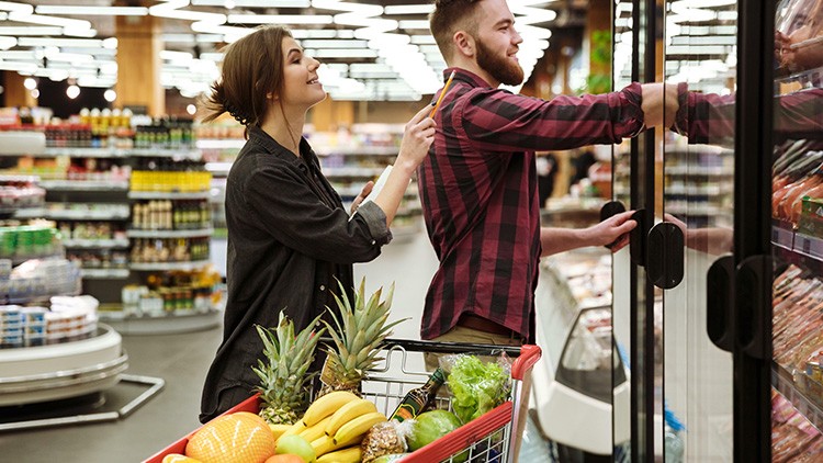Einkaufswagen mit Obst und Gemüse steht vor einer Kühltheke, zwei Personen greifen nach einem Produkt aus der Kühlung.