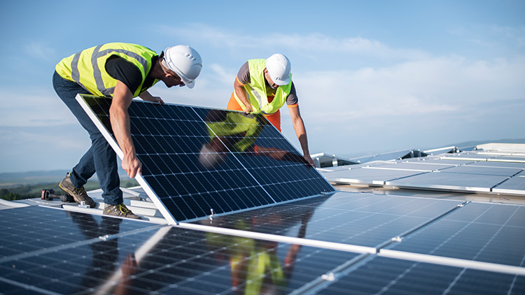 Zwei Personen in Warnwesten und Schutzhelmen installieren ein Solarpanel auf einem Dach bei klarem Himmel.