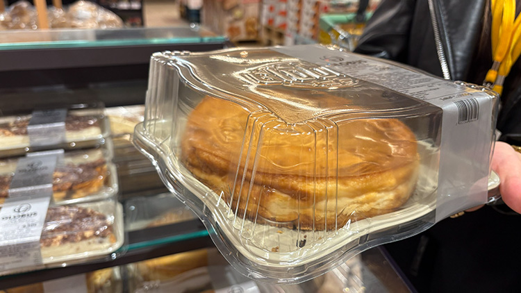 Eine goldbraune, verpackte Käsekuchenportion wird von einer Hand gehalten, daneben liegt Gebäck in Scheiben.