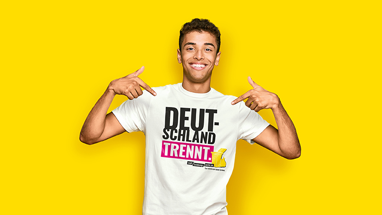 Person in weißem T-Shirt mit Aufschrift „DEUTSCHLAND TRENNT“, gelbem Symbol und rosa Akzent vor gelbem Hintergrund zeigt auf sich.