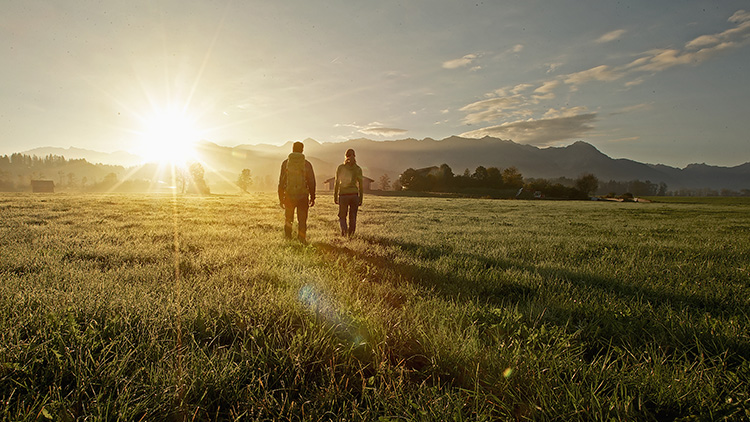 Zwei Personen mit Rucksäcken gehen über eine Wiese in Richtung Sonnenaufgang, im Hintergrund Berge und Bäume.