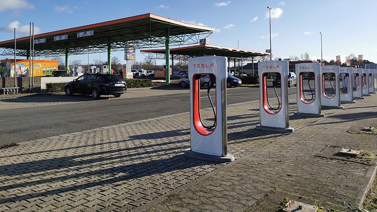 Mehrere Tesla-Ladesäulen stehen auf einem gepflasterten Parkplatz neben einer überdachten Tankstelle bei Sonnenschein.